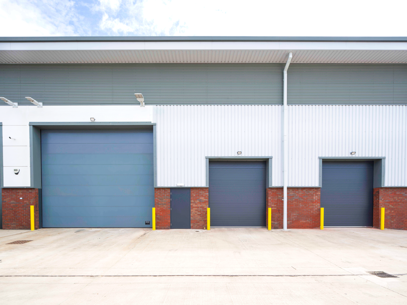 Industrial warehouse exterior featuring three grey roller shutters, brick and metal cladding, yellow bollards, and a concrete loading bay.
