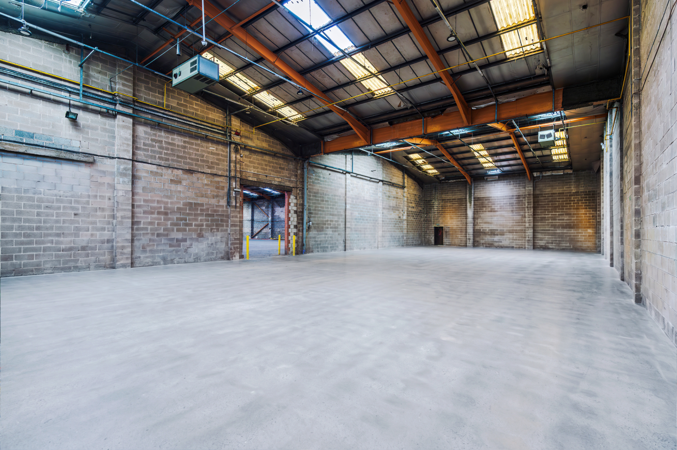 Large, empty industrial warehouse with concrete floor, exposed brick walls, high ceilings, rooflights, and industrial lighting.