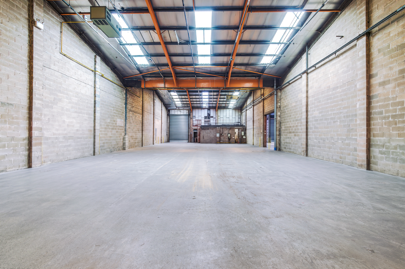 Empty industrial warehouse with concrete floor, brick walls, and exposed steel beams. Large roller shutters, skylights let in light.