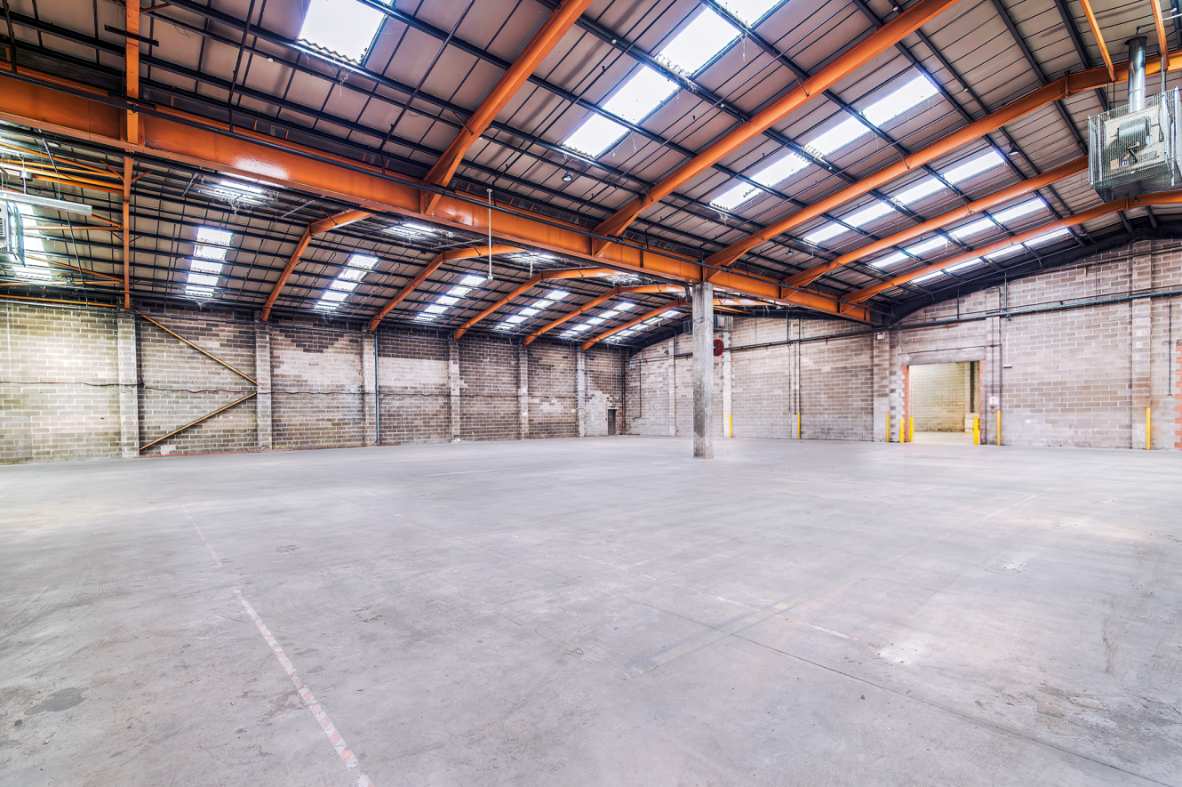 Empty industrial warehouse with high ceilings, skylights, exposed beams, concrete floor, brick walls, and loading bay door visible.
