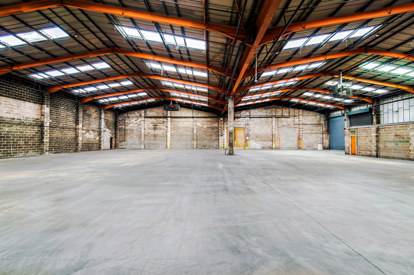 Large empty warehouse interior with high ceilings, exposed brick walls, metal beams, concrete floor, and skylights above.