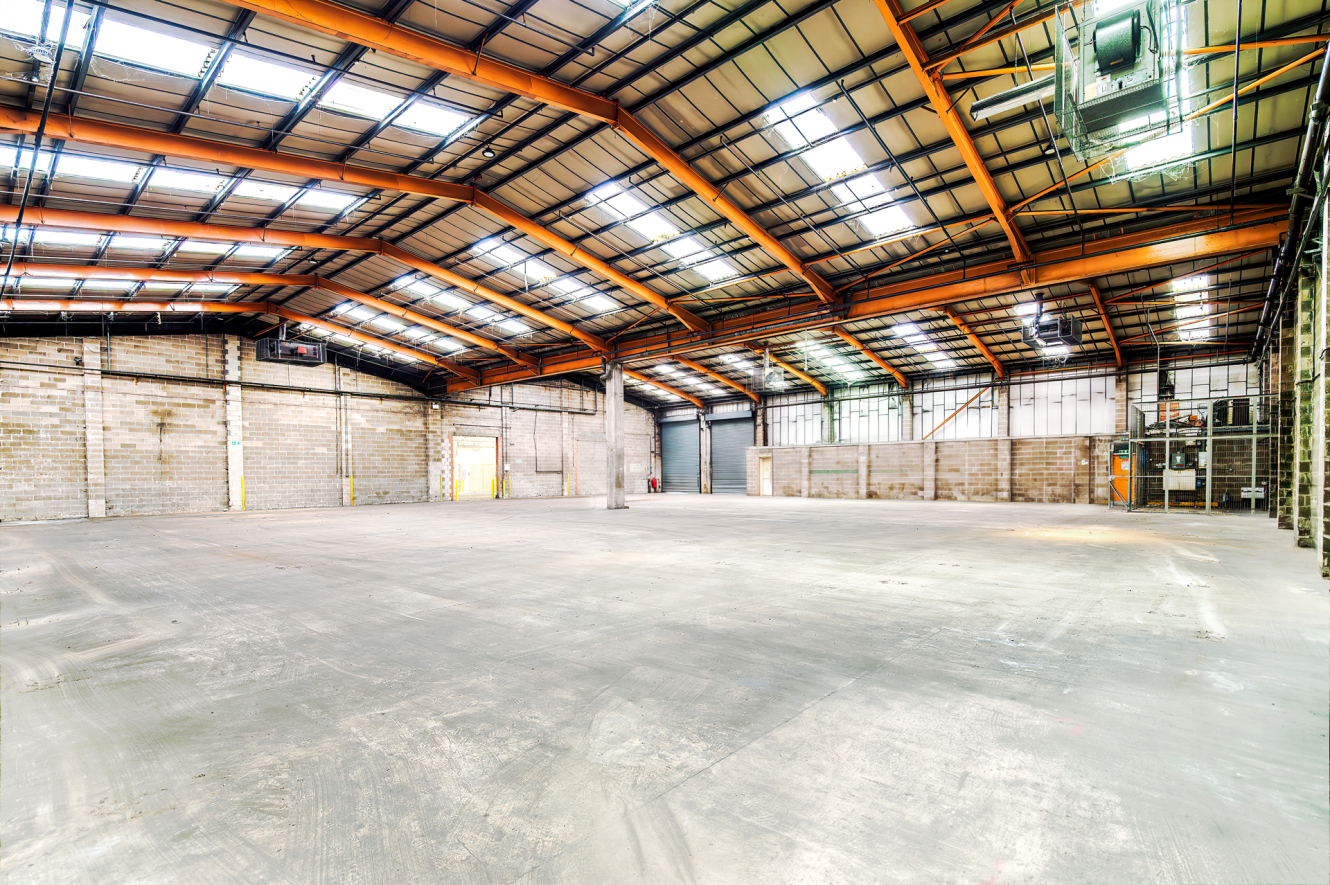 Large empty industrial warehouse with high ceilings, exposed beams, concrete floors, brick walls, and skylights letting in sunlight.
