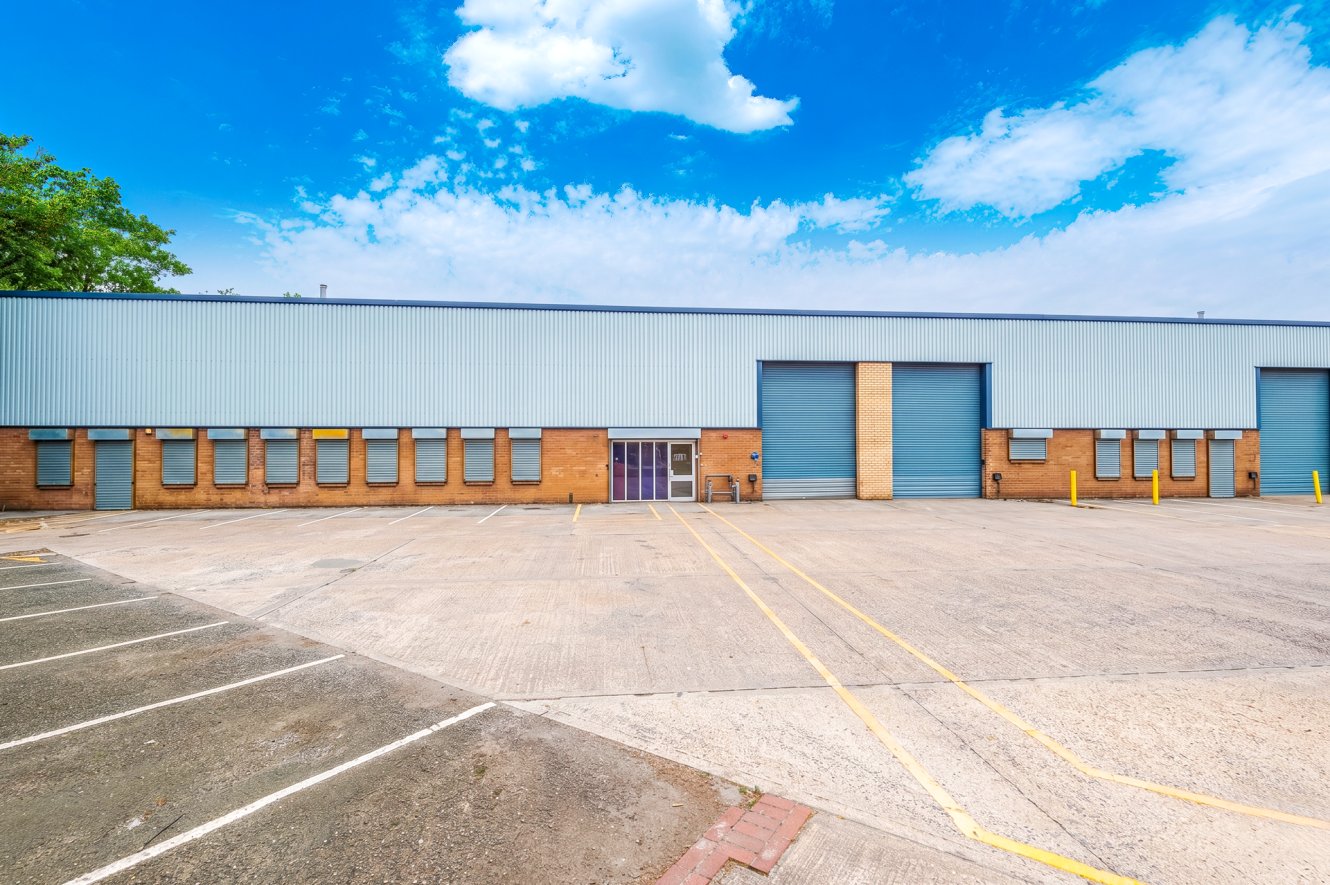 A wide, empty car park in front of a warehouse with blue roller doors, brick and metal cladding, and a glass entrance.