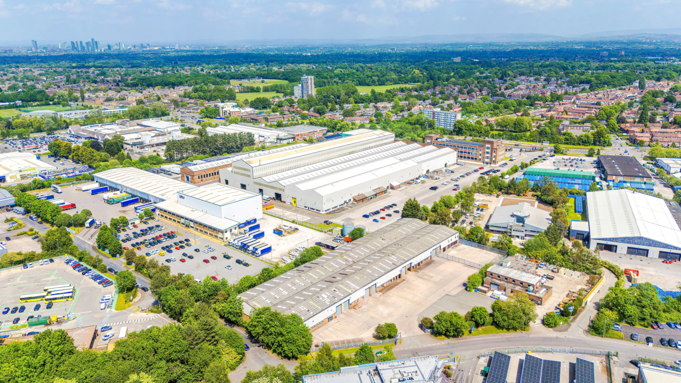 Aerial view of an industrial area with large warehouses, car parks, roads, greenery, and distant residential buildings.