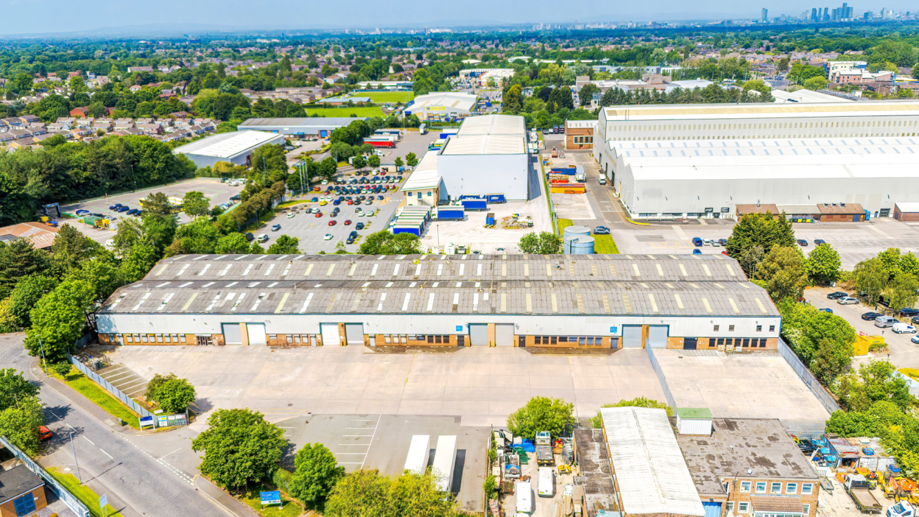 A large industrial warehouse complex with loading bays, car parks, trees, and nearby buildings under a clear sky.