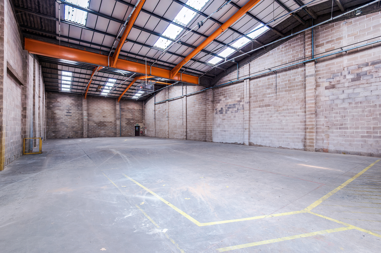 Empty industrial warehouse with concrete floors, high ceilings, exposed beams, brick walls, and overhead rooflights. Yellow floor markings.