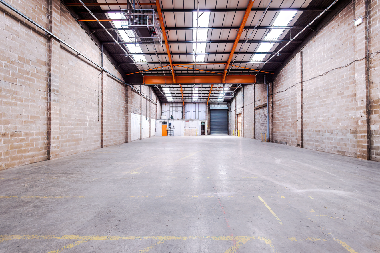 Interior of an empty industrial warehouse with high ceilings, exposed brick walls, concrete floor, and large roller shutters at the far end.
