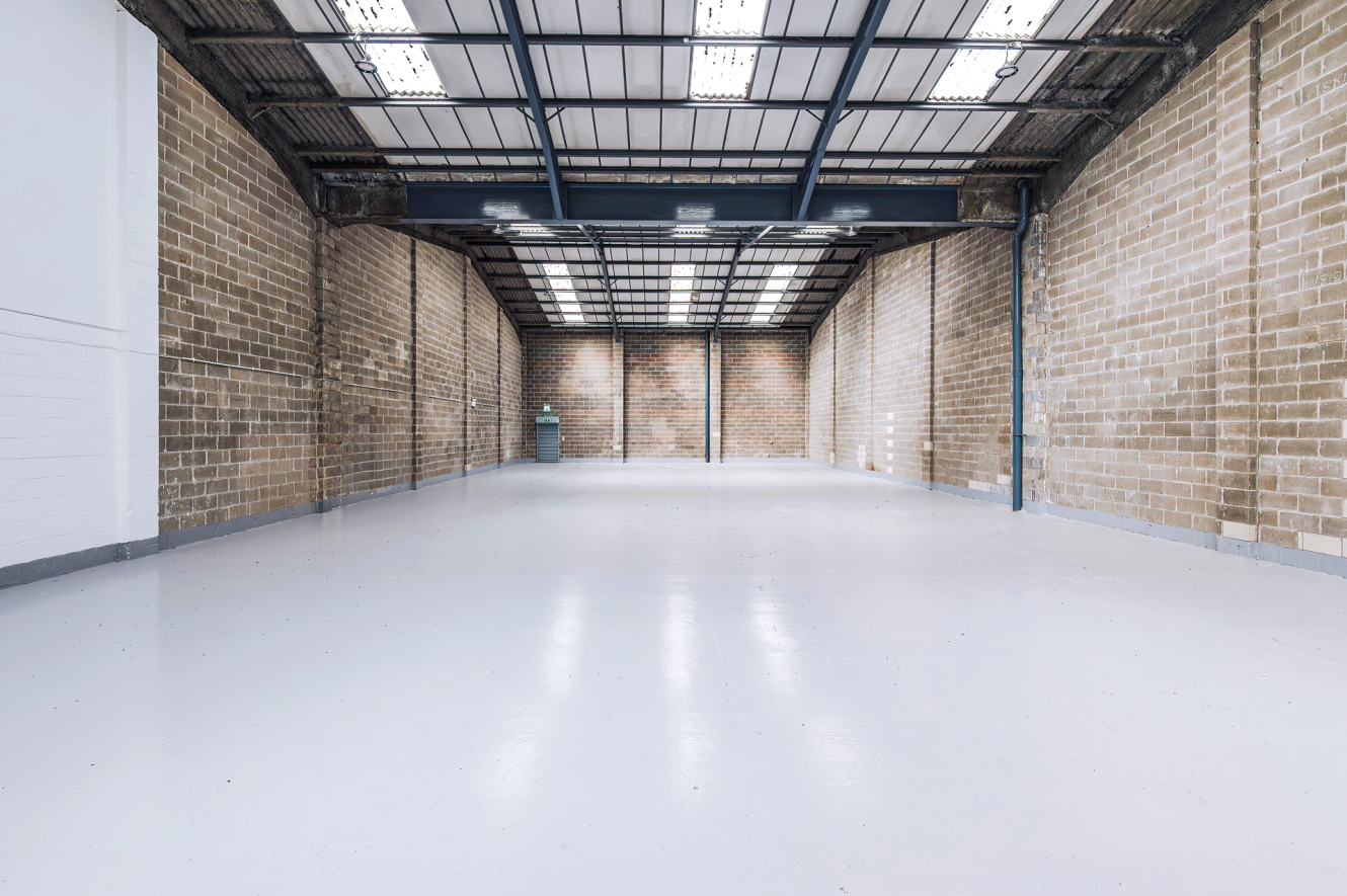 Empty warehouse with brick walls, high ceiling, exposed beams, fluorescent lights, and a polished concrete floor.