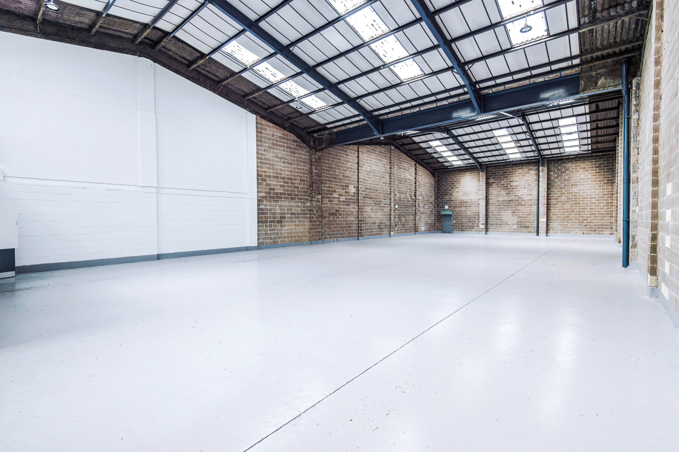 Empty warehouse interior featuring high ceilings, exposed brick walls, metal beams, and a polished concrete floor under bright lighting.