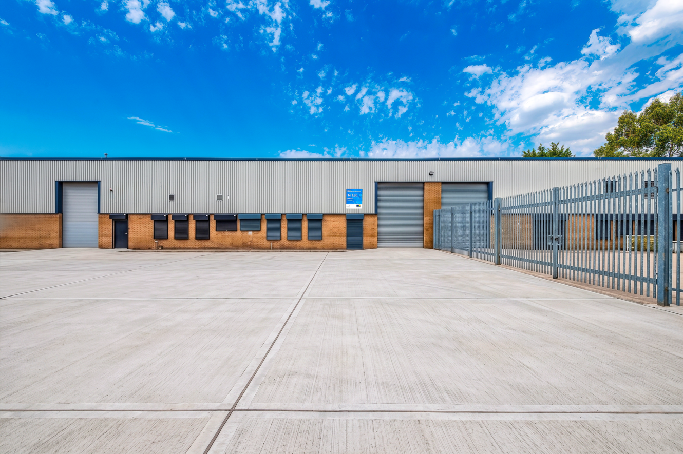 Industrial warehouse featuring roller shutters, brick and metal cladding, and a fenced concrete yard beneath a blue sky with clouds.
