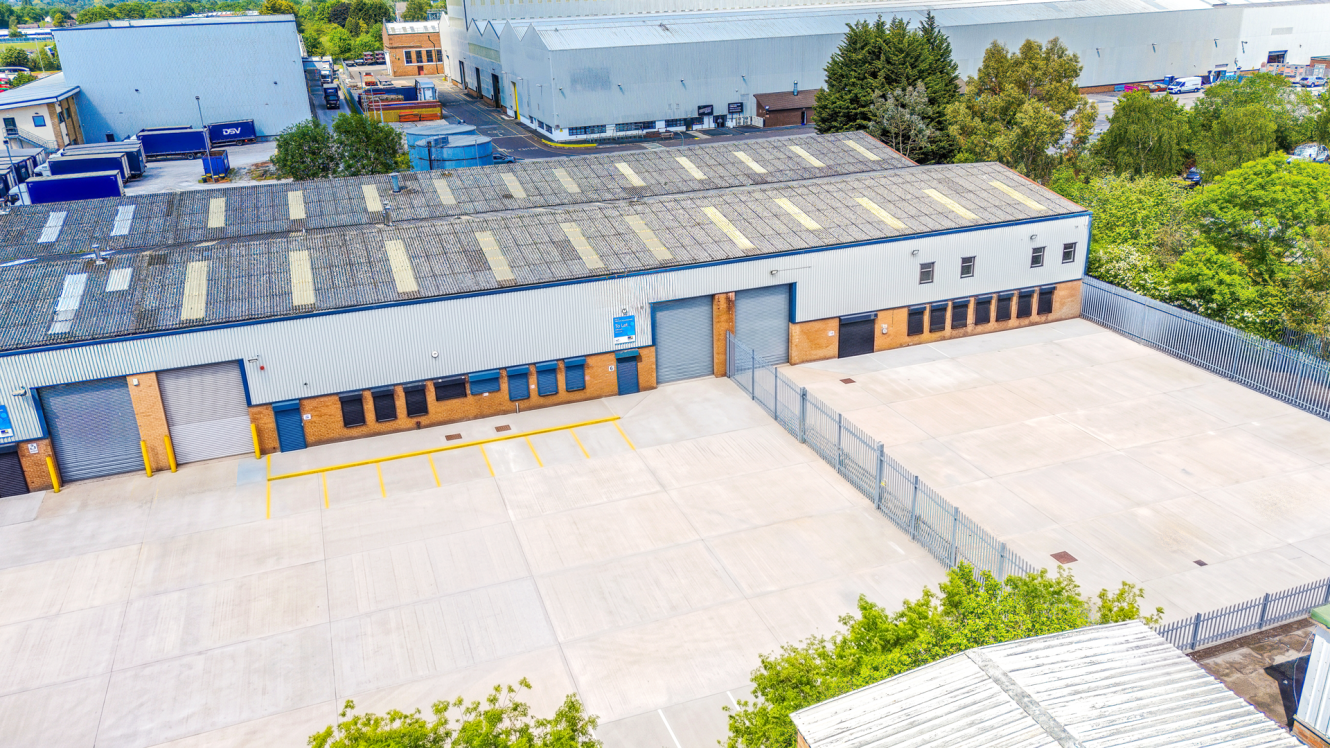 Aerial view of a large industrial warehouse with a gated, empty concrete car park and greenery surrounding the building.