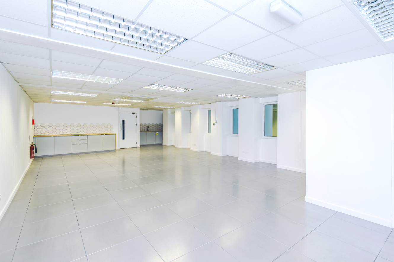 Spacious, empty office room with tiled flooring, white walls, ceiling lights, windows, and a kitchenette area on one side.