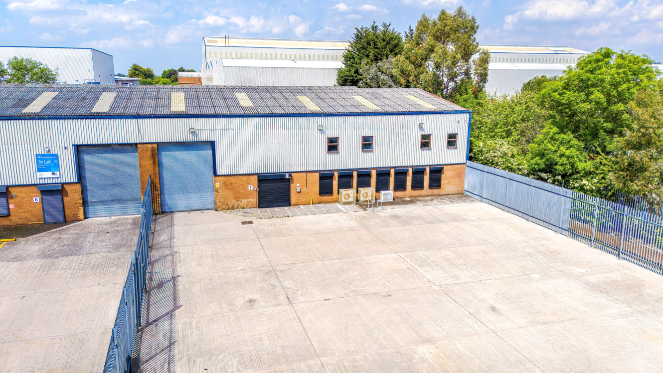 Industrial warehouse with blue roller doors, corrugated metal cladding, fenced yard, windows, air conditioning units. Trees and buildings behind.