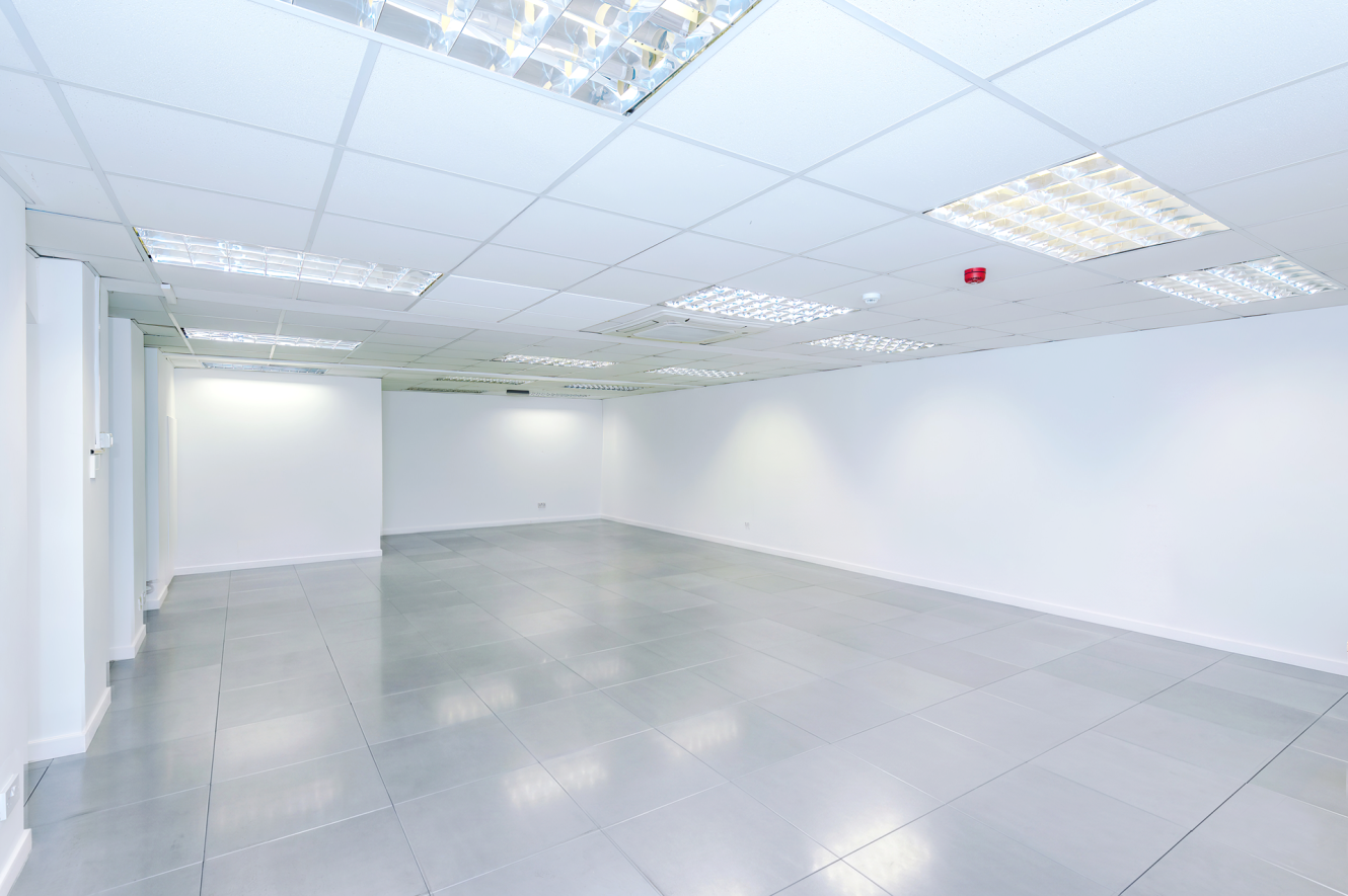Empty, brightly lit office space featuring white walls, a grey tiled floor, and a suspended ceiling fitted with fluorescent lights.