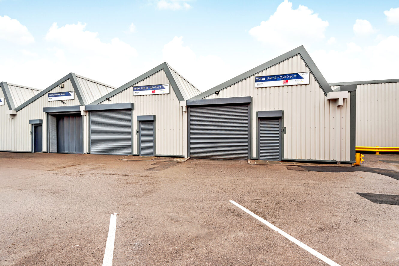 A row of industrial warehouse units with metal roller shutter doors stands on an empty tarmac car park under a partly cloudy sky.
