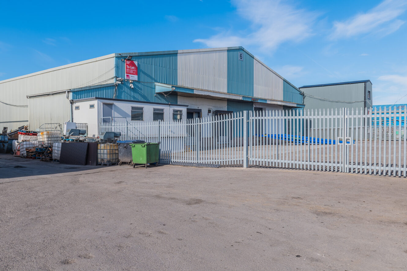 A large industrial warehouse with a metal fence, scattered equipment, and skips outside beneath a clear blue sky.