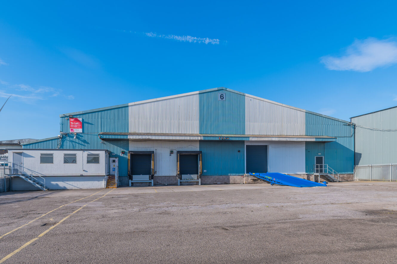 Exterior view of a large warehouse with three loading bays, a ramp, and a small office on the left beneath a clear blue sky.