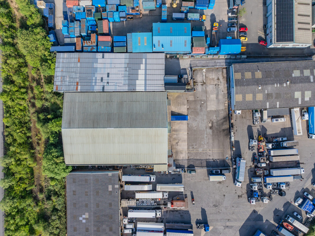 Aerial view of an industrial area with warehouses, lorries, shipping containers, and green vegetation on the left side.