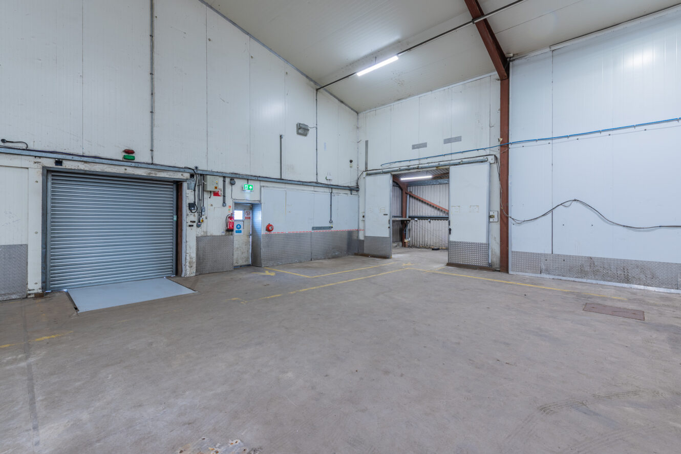 Empty industrial warehouse with a concrete floor, two large loading doors, white walls, and overhead lighting.
