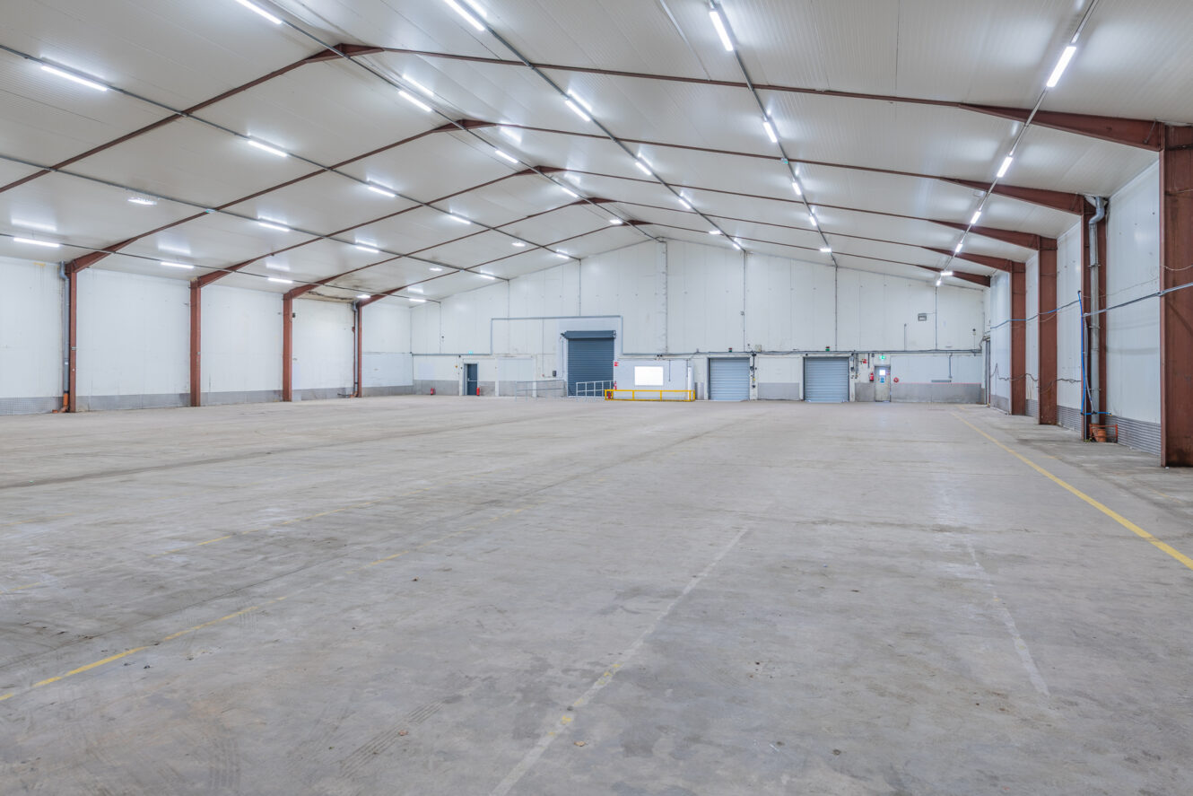 Interior of a large, empty warehouse with high ceilings, steel beams, concrete floor, and multiple loading bay doors at the back.