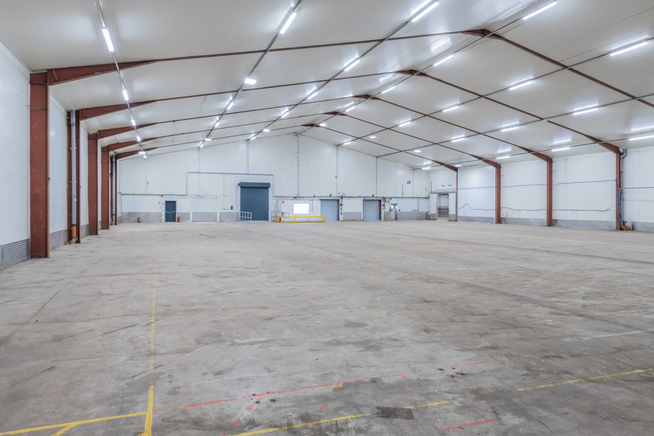 Spacious, empty warehouse interior with high ceiling, exposed steel beams, large doors, and bright lighting. Concrete floor marked.
