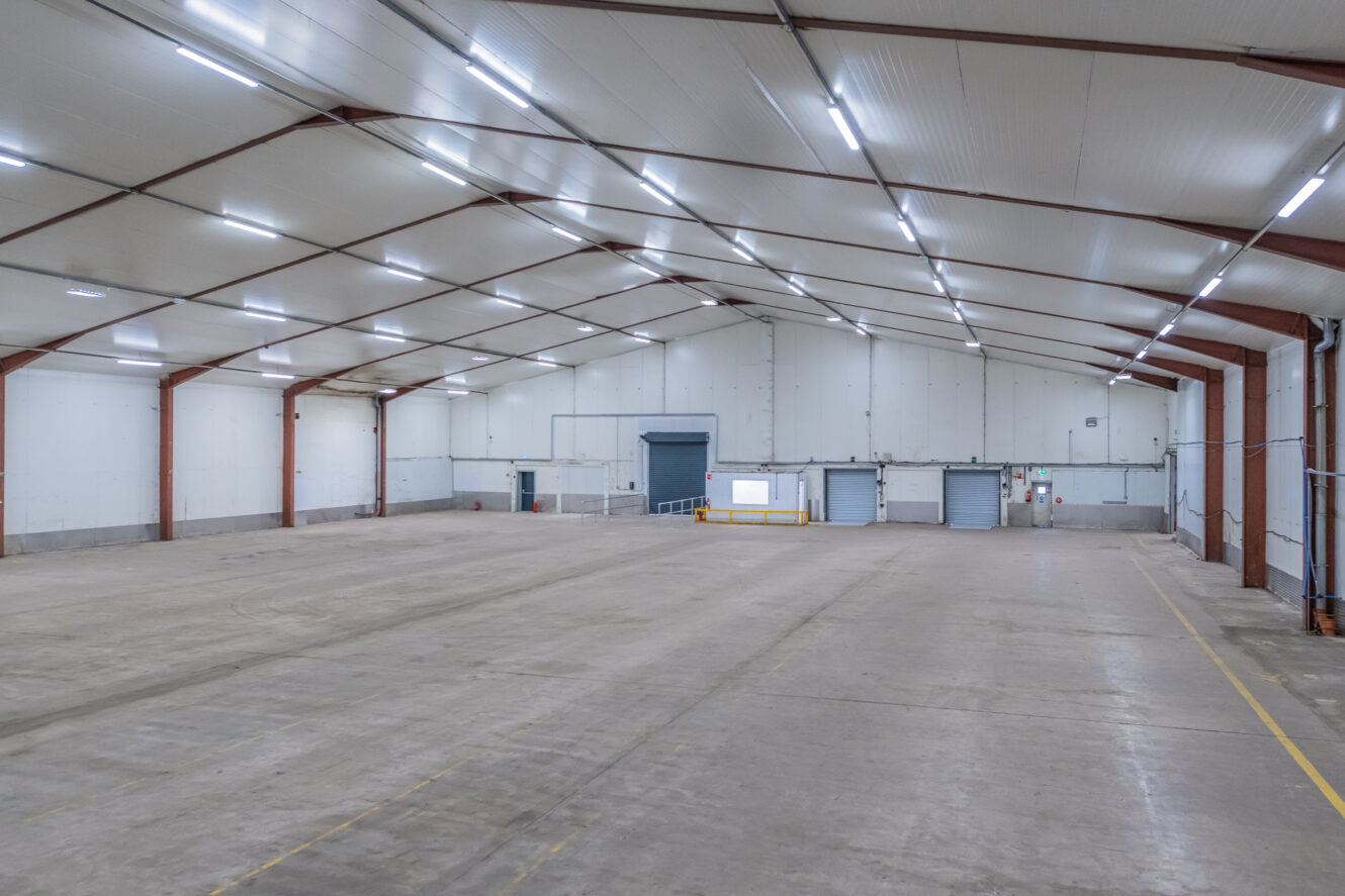 Spacious, empty warehouse with high ceilings, white walls, several loading bay doors, and overhead lighting.
