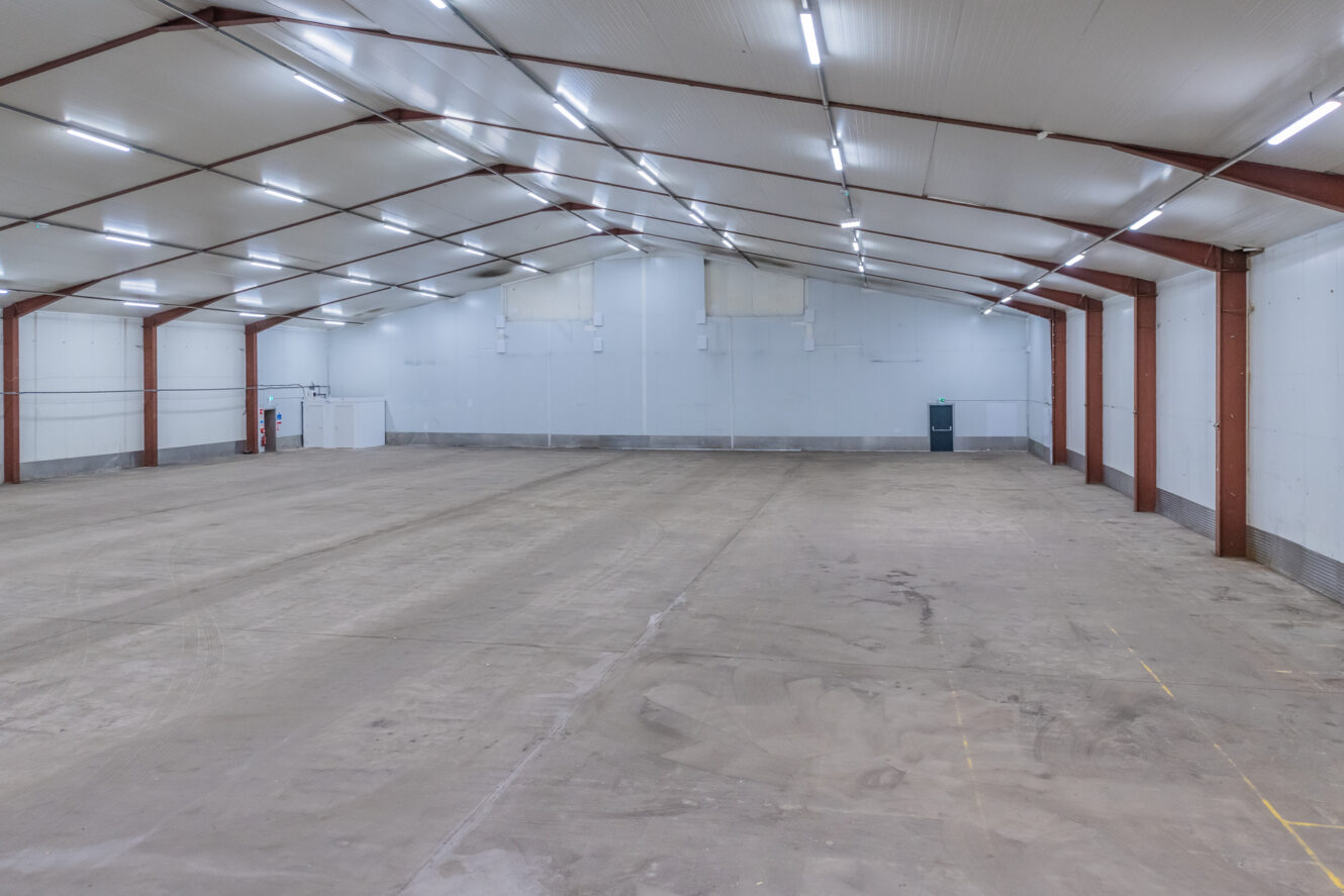 Empty warehouse interior with high ceiling, white walls, red supports, bright lighting, concrete floor; extinguisher and bin on wall.