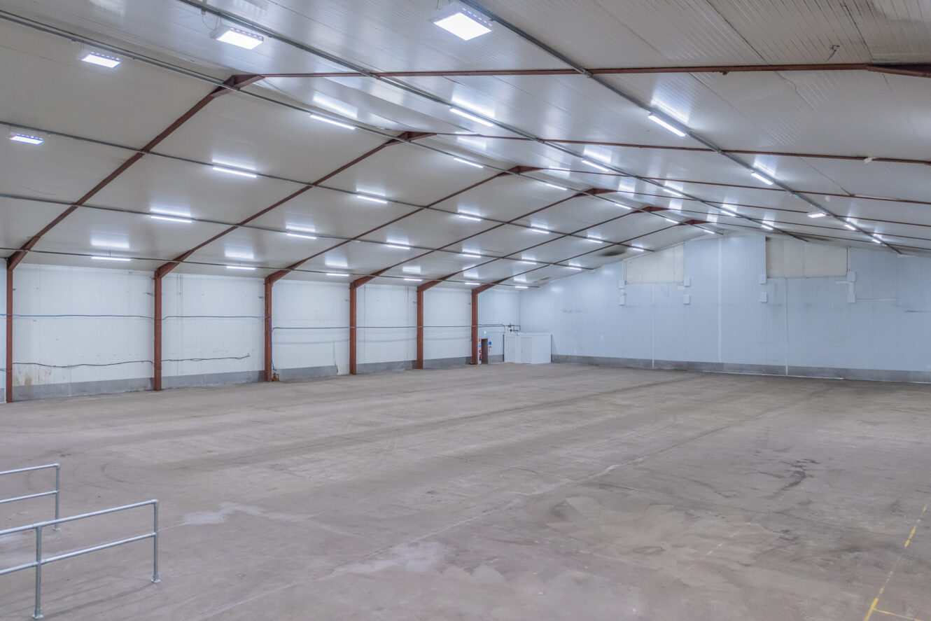 Large, empty warehouse interior with high ceilings, white walls, exposed beams, bright overhead lights, and a concrete floor.