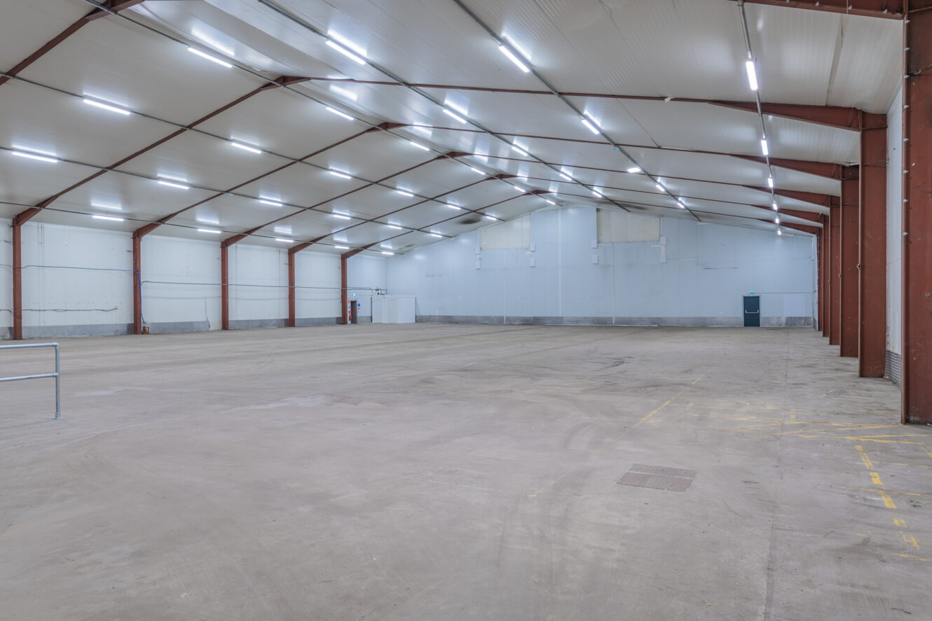 Large empty warehouse interior with high ceiling, exposed red steel beams, concrete floor, and bright overhead lights.