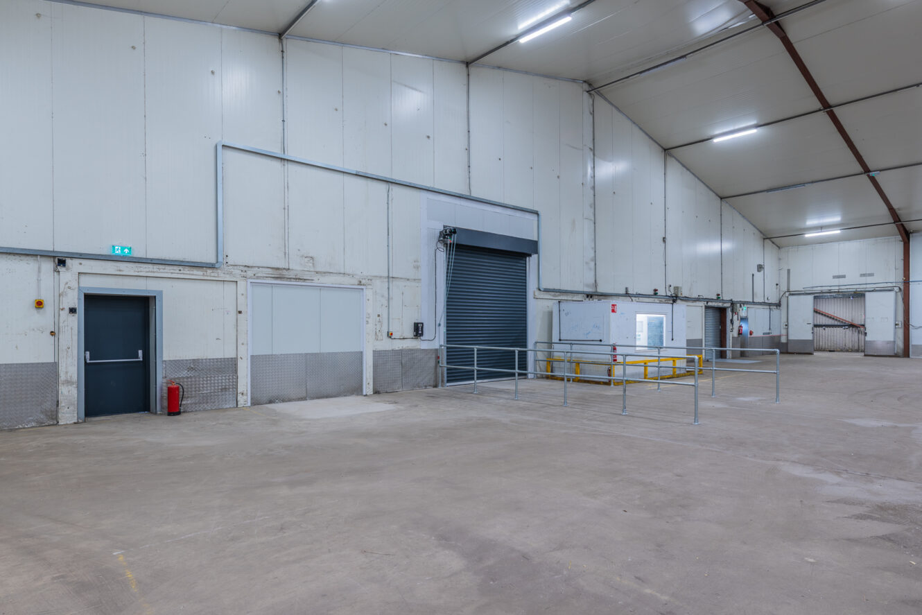 Interior of a vacant industrial warehouse featuring a closed loading bay door, safety railings, fire extinguisher, and high ceilings.