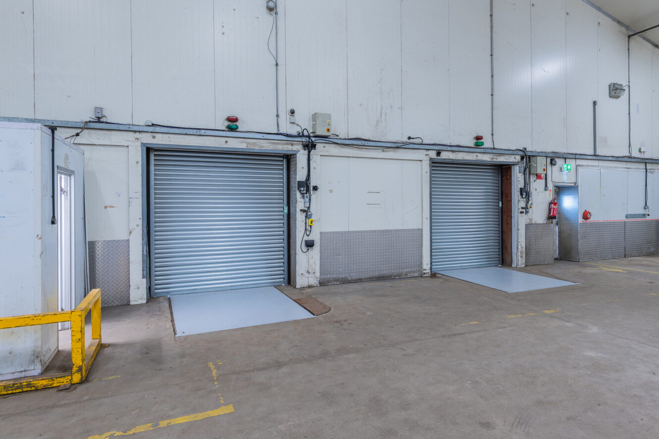 Three closed roller shutter doors in a warehouse with concrete floors and white walls; safety and control equipment are present.