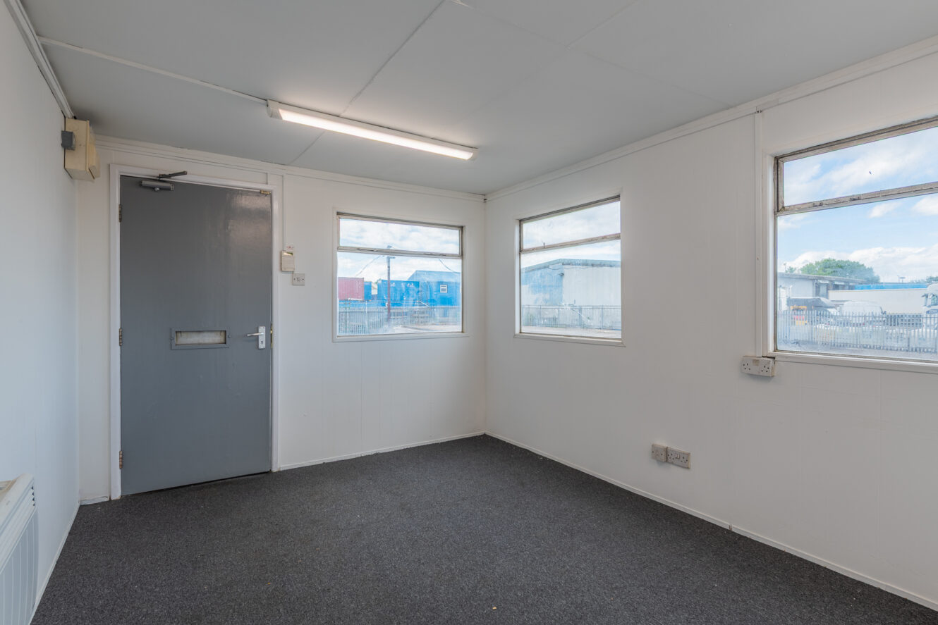 An empty room with grey carpet, white walls, a grey door, and two windows revealing an industrial area. The ceiling light is on.