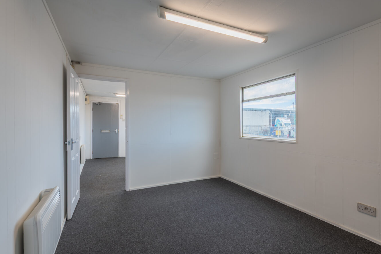 Empty office room with white walls, grey carpet, window, fluorescent ceiling light, and open door leading to a corridor.