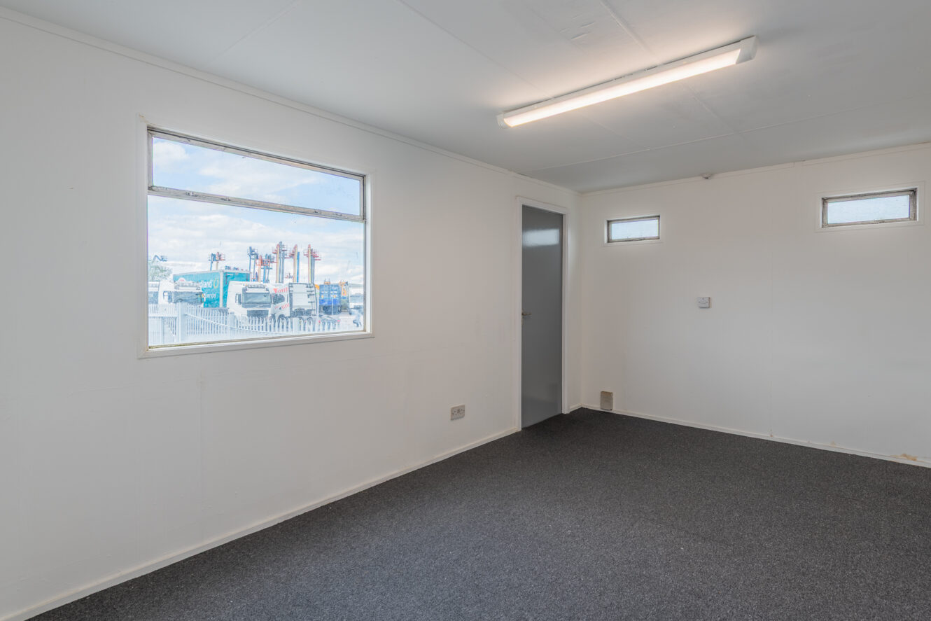 Unfurnished room with white walls, grey carpet, fluorescent ceiling light, and three windows; door opens to another room. Industrial area seen outside main window.