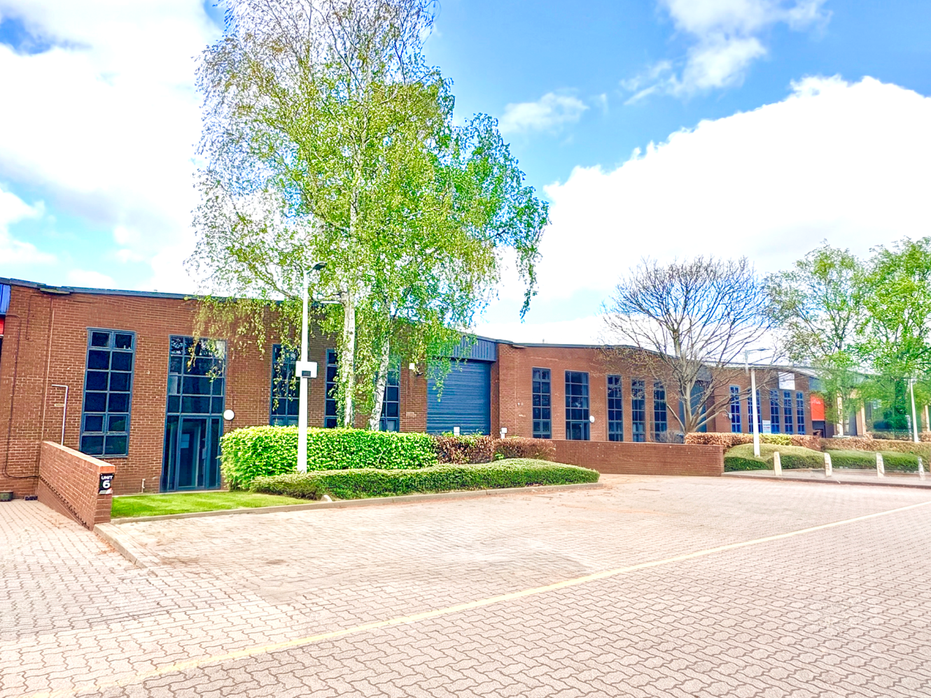 Single-storey brick office building with tall vertical windows, trees and bushes around it, next to a paved car park under cloudy skies.