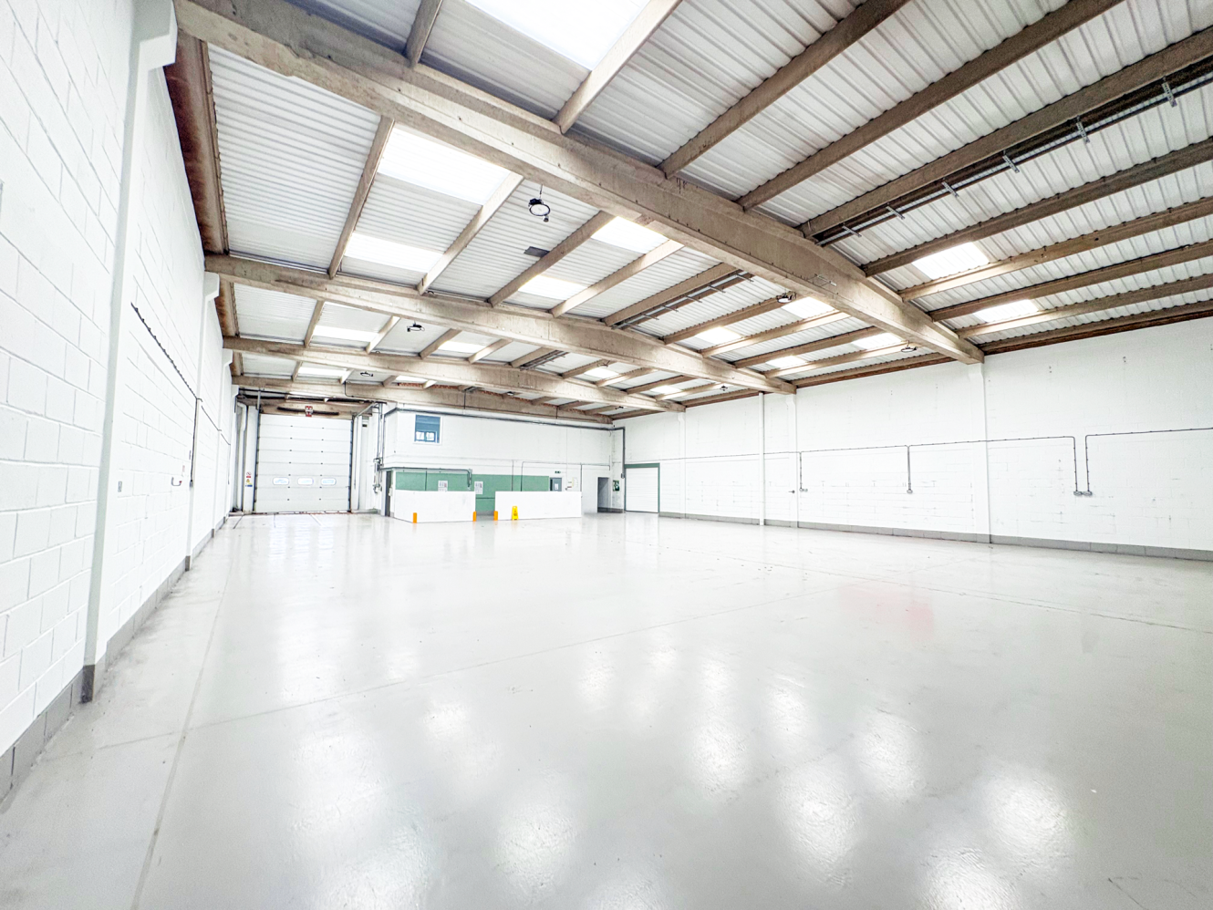 Large empty warehouse with white walls, polished concrete floor, open beams, ceiling skylights, doors and a loading bay at the end.