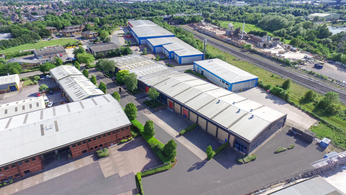 Aerial view of an industrial estate with warehouses, roads, parked vehicles, railway tracks, and greenery nearby.