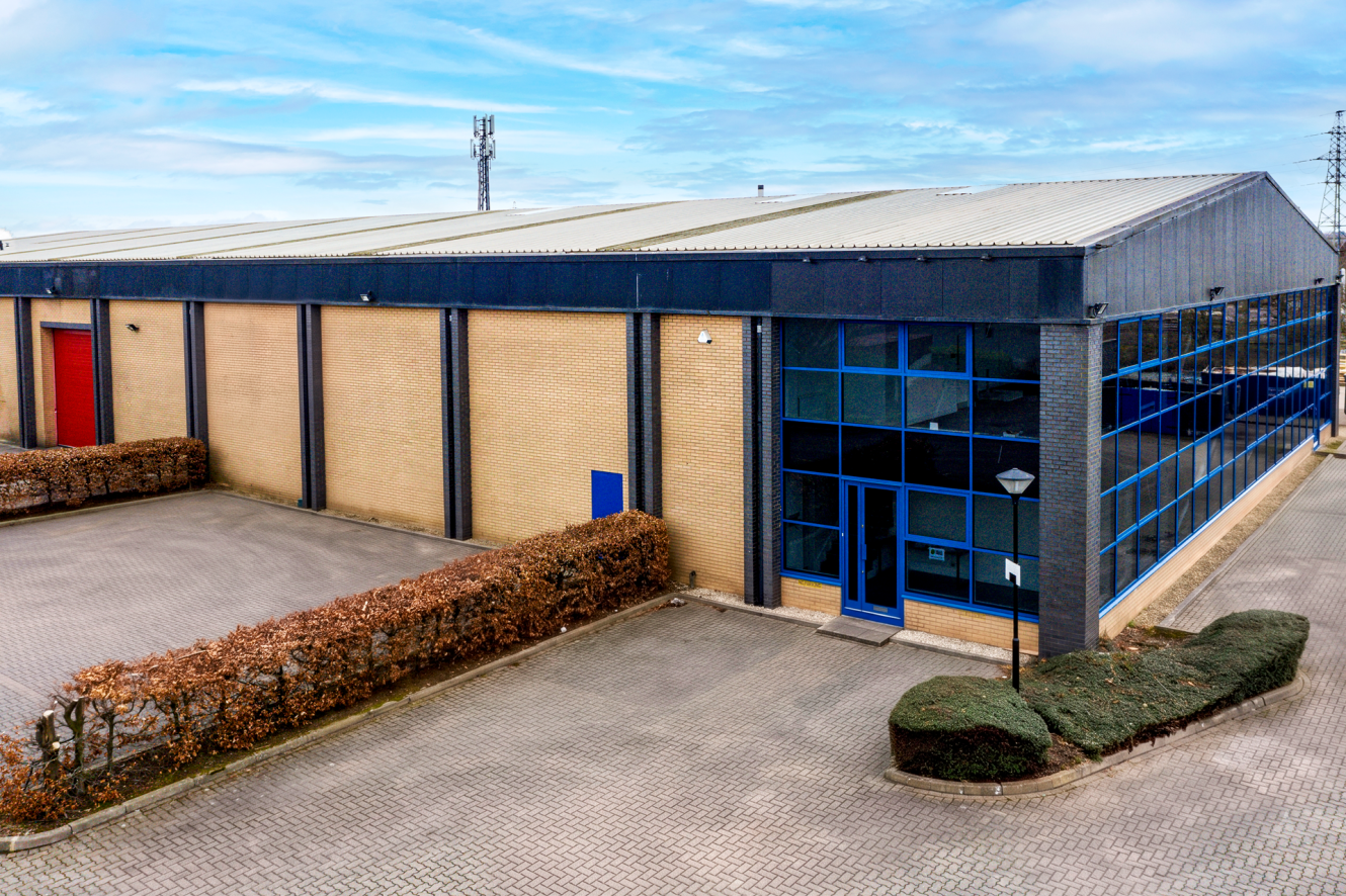A modern industrial warehouse with large glass windows, brick walls, and a vacant tarmacked car park under a partly cloudy sky.