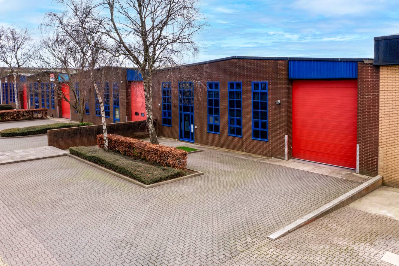 Industrial warehouse units with red roller shutters and blue-framed windows, bare trees in front, on a paved car park under a partly cloudy sky.
