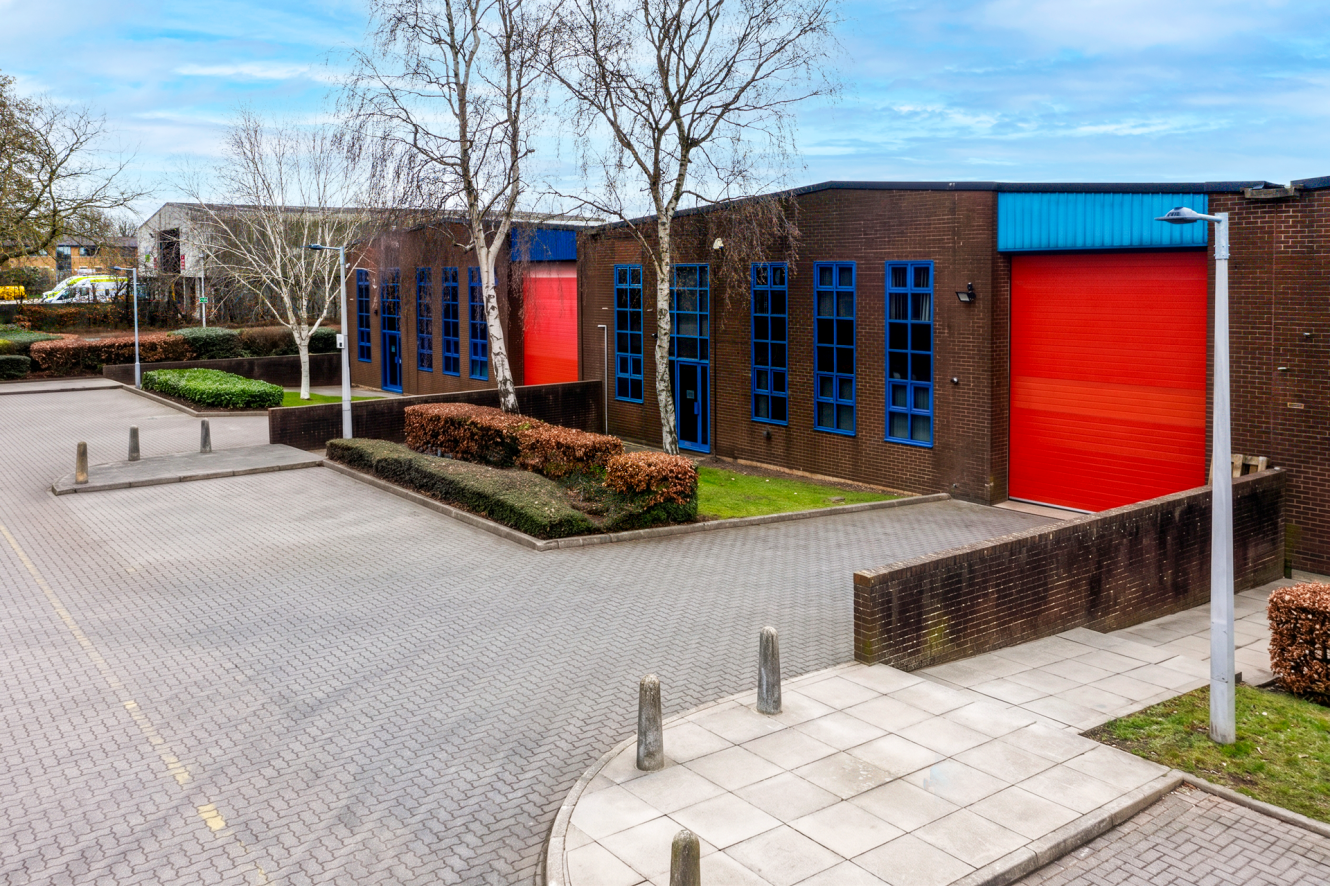 Modern industrial building with red roller shutters and blue-framed windows, surrounded by paved areas and landscaped greenery.