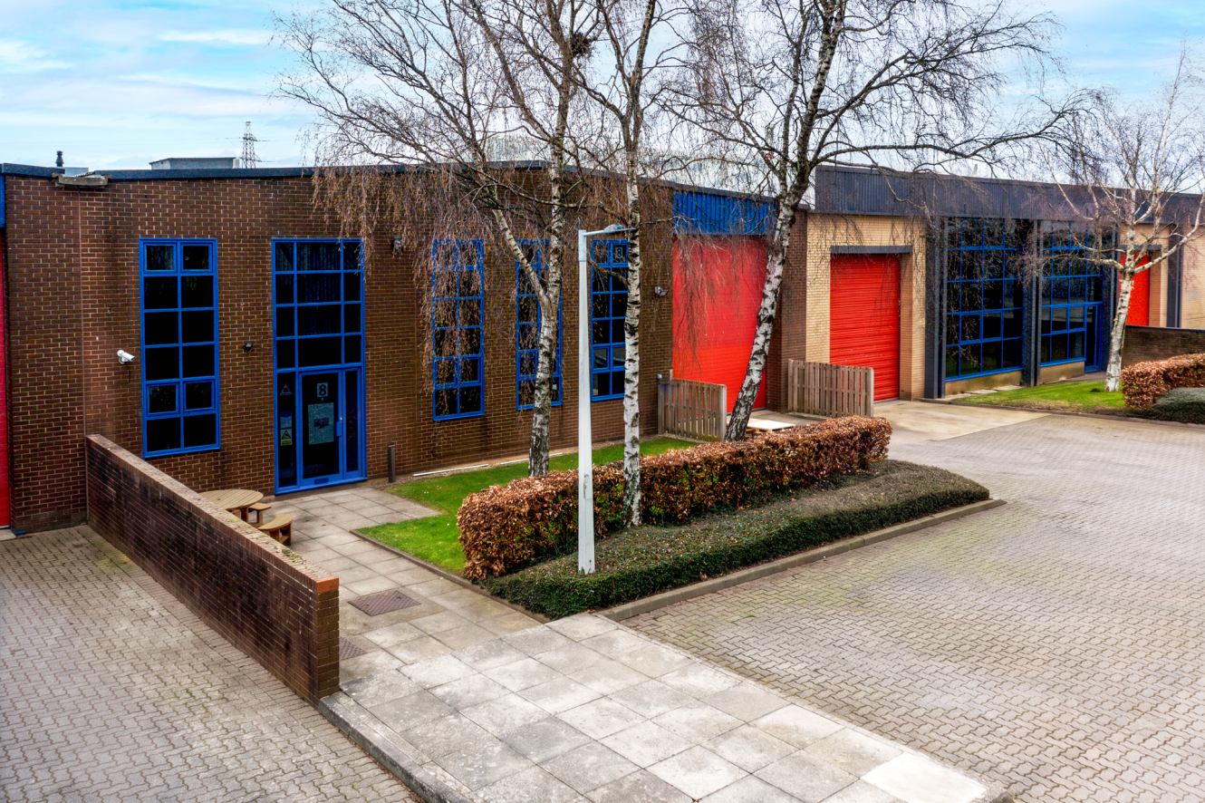 A row of industrial buildings with brick walls, blue window frames, red roller shutters, and bare trees by a paved drive and walkway.