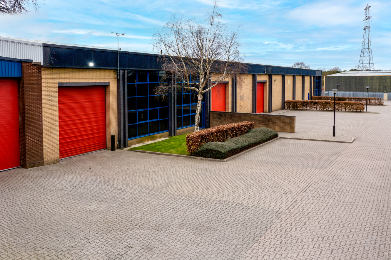 Industrial buildings of brick and blue-tinted glass, several red roller shutters, a tree, trimmed hedges, and an empty paved car park.
