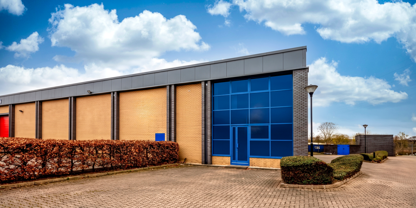 Modern industrial warehouse with beige brick walls, blue glass windows, and an empty paved car park under a partly cloudy sky.