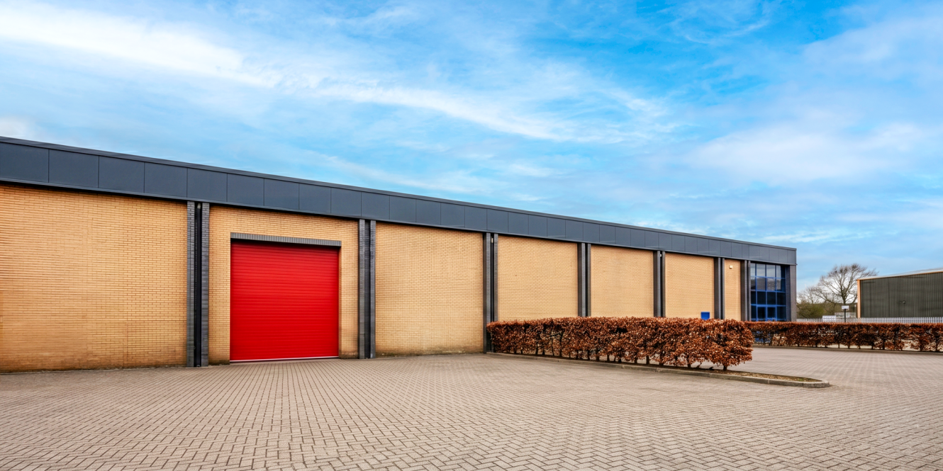 A modern industrial warehouse with a brick exterior, red roller shutter door, blue-tinted windows, and paved parking under a cloudy sky.