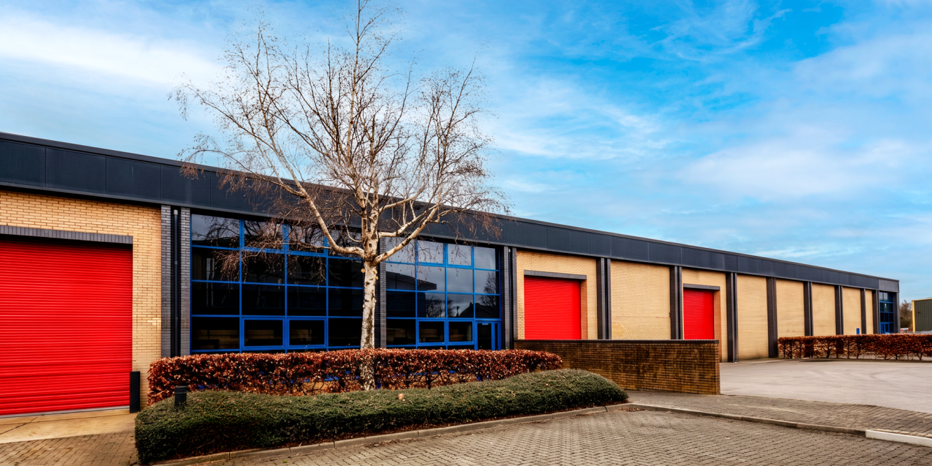 A modern industrial warehouse with red roller shutters, blue windows, bare tree, trimmed hedges in front, under partly cloudy sky.