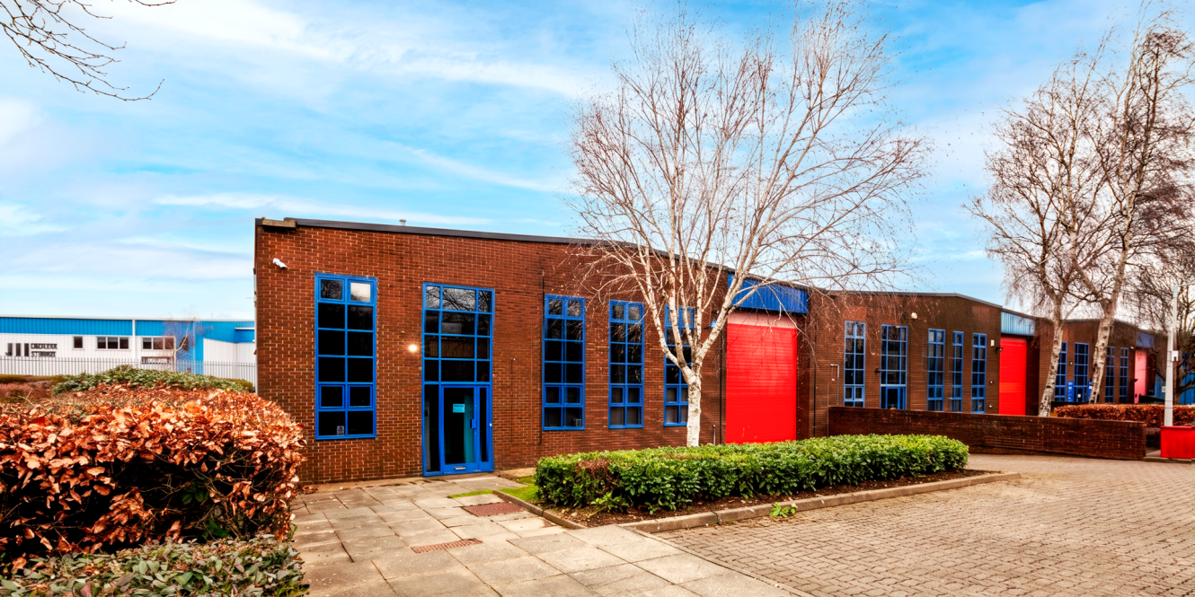 Row of modern industrial units with brown brick walls, blue-framed windows, and large red doors, surrounded by bushes and trees.