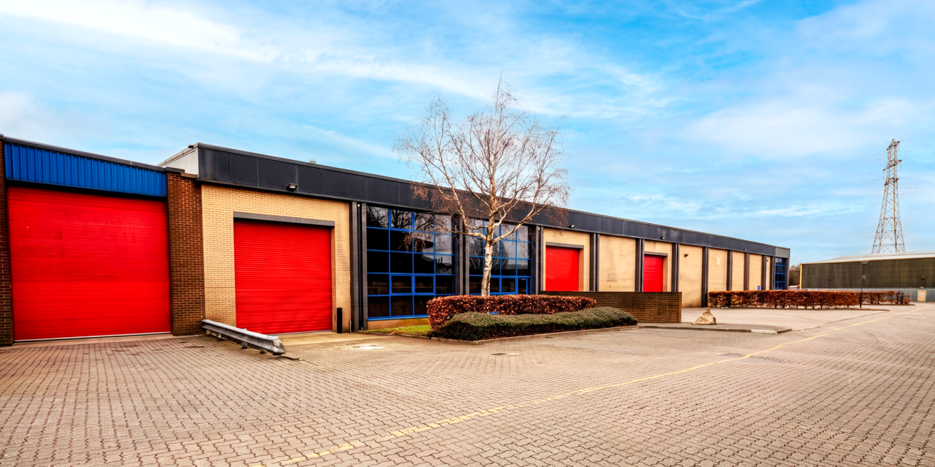 A row of industrial warehouse units with red roller shutters, large windows, and a paved driveway beneath a blue sky.