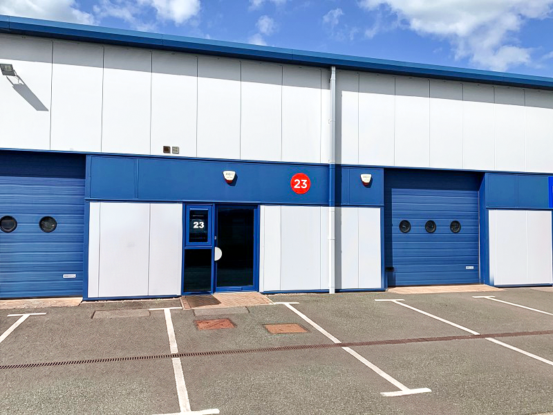 Exterior of a blue and white industrial unit with three doors, number 23 above the entrance and on a door, empty car park in front.