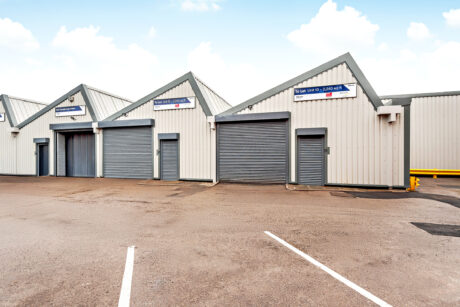 A row of industrial units with closed grey roller shutter doors, white and grey cladding, and empty parking spaces in front.