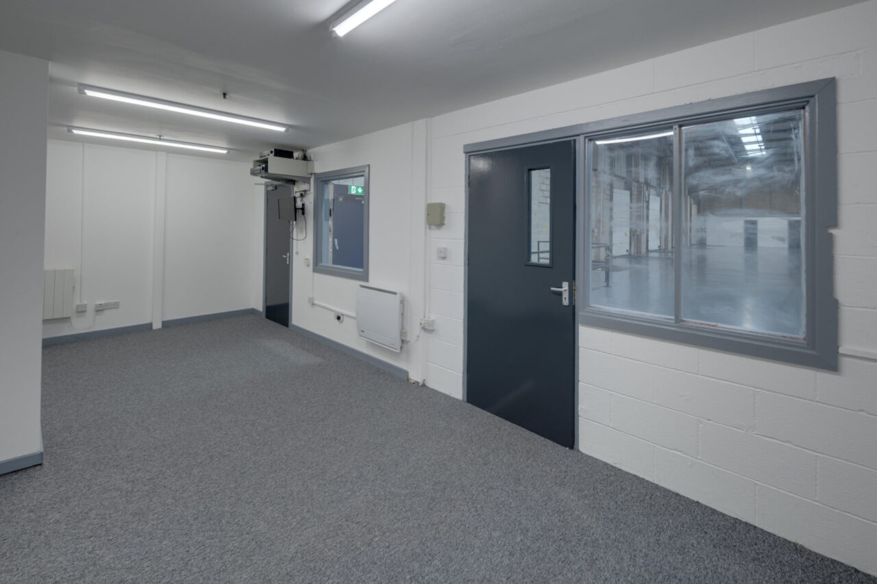 Empty office with grey carpet, white walls, ceiling lights, and grey doors; a window overlooks an adjoining industrial area.